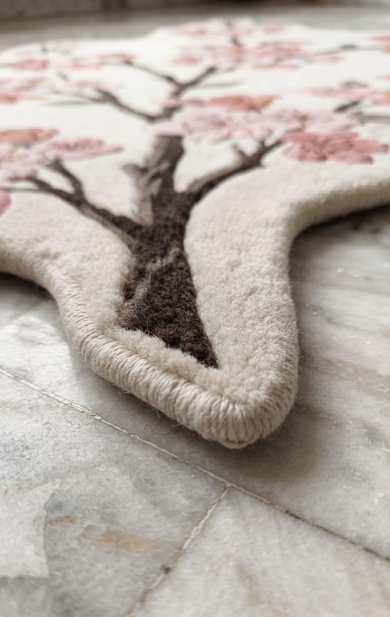  Detailed-3d-hand-carved-high-low-pile-texture-new-zealand-wool-pink-sakura-flower-rug A close-up of a plush rug featuring a stylized cherry blossom tree design in cream, pink, and dark brown, laid on a light marble floor.