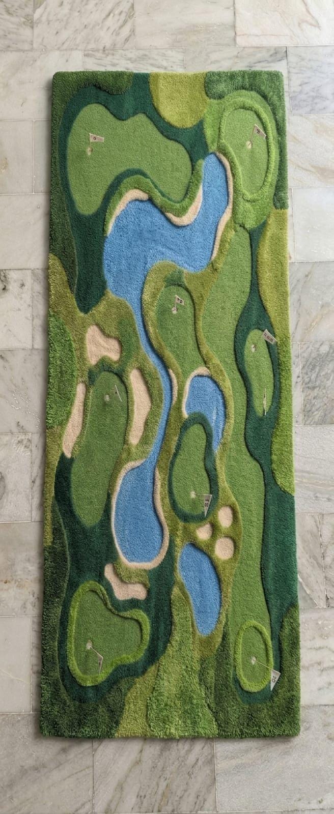 Modern-sports-lounge-interior-styling-featuring-luxury-green-golf-course-shaped-accent-carpet A rectangular plush rug featuring a stylized golf course landscape with green fairways, blue water hazards, and beige sand bunkers, displayed on a light gray marble tile floor.