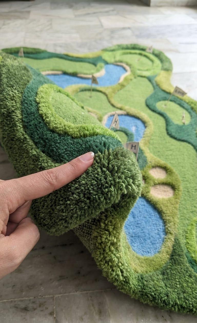 Detailed-3d-topography-hand-carved-high-low-pile-texture-fairway-green-wool-rug A close-up of a green golf course themed rug being held by a hand, showing its high-pile texture and raised terrain details with small flagstick markers.