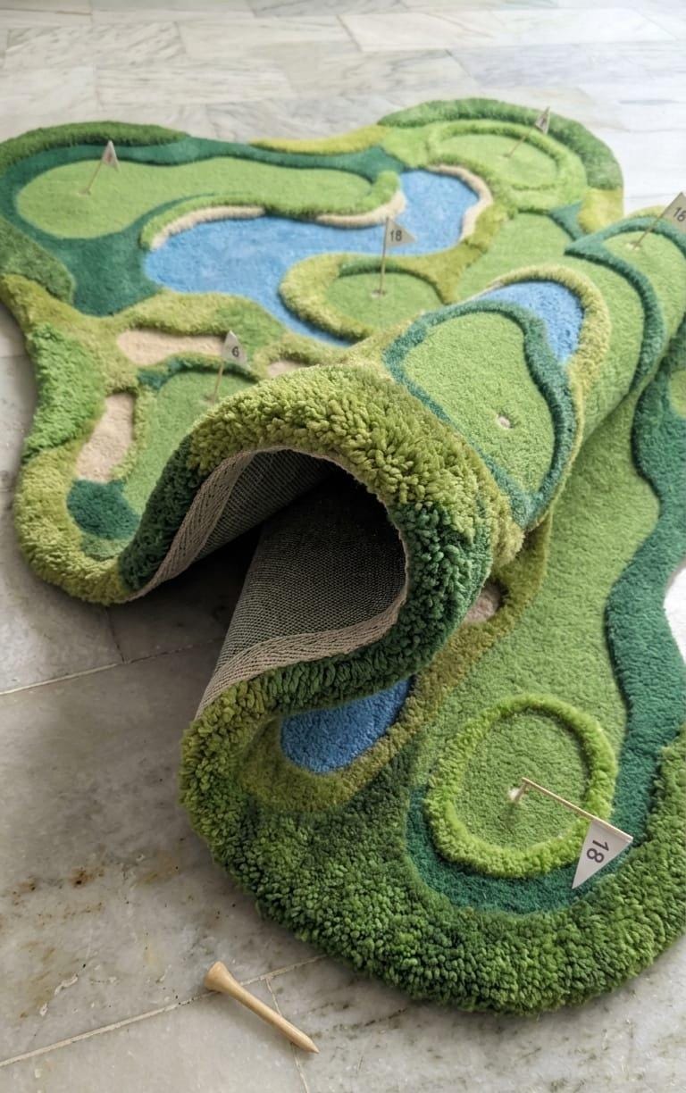 Detailed-3d-topography-hand-carved-high-low-pile-texture-fairway-green-wool-rug A novelty golf course rug with raised green grass textures, blue water features, and sand bunkers, shown partially rolled up on a light stone floor.