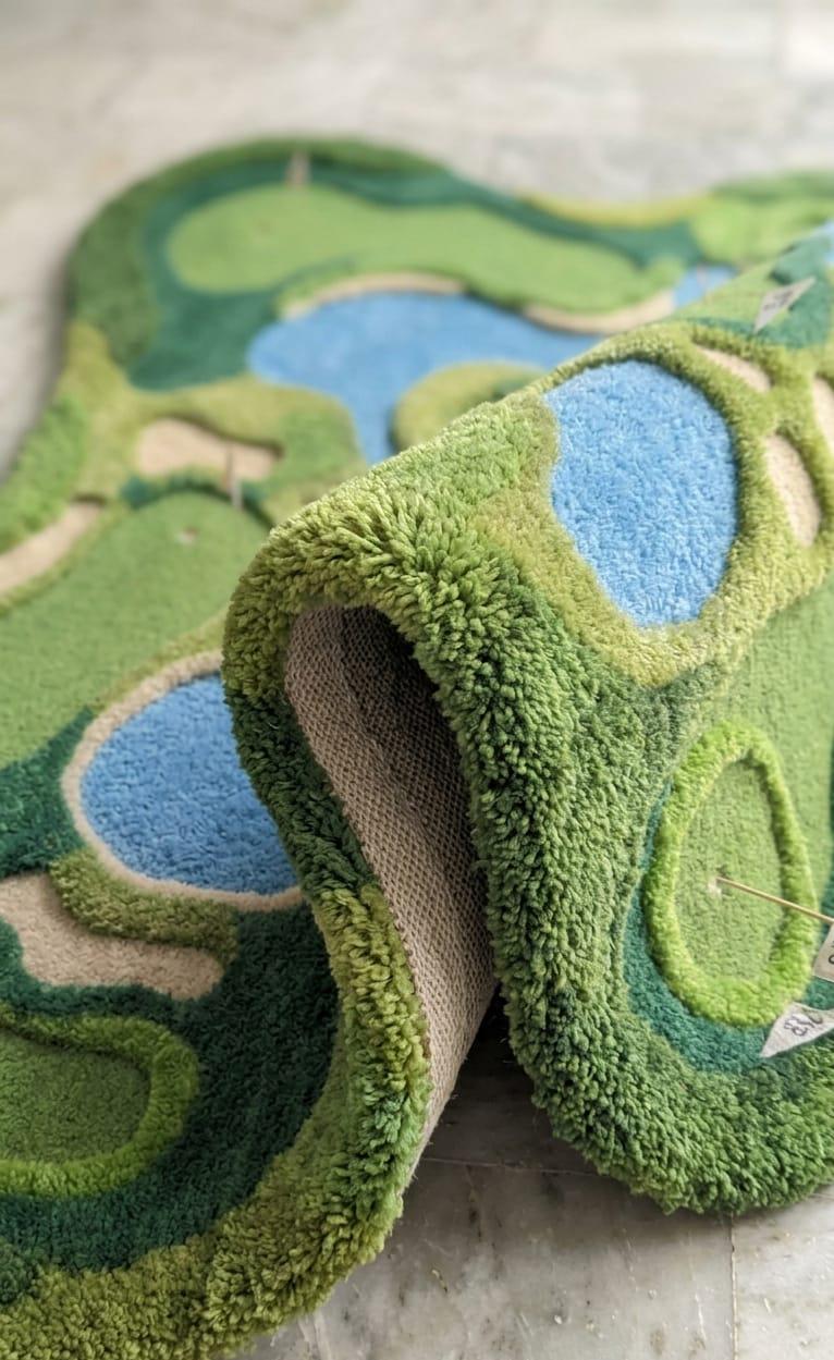 Shubham-Art-Rugs-hand-tufted-golf-course-rug-new-zealand-wool-3d-topography-sports-room-decorDetailed-3d-topography-hand-carved-high-low-pile-texture-fairway-green-wool-rug A close-up of a plush, sculptural rug with a golf course design, featuring green and blue tufted patterns on a textured beige backing, shown on a light gray stone floor.