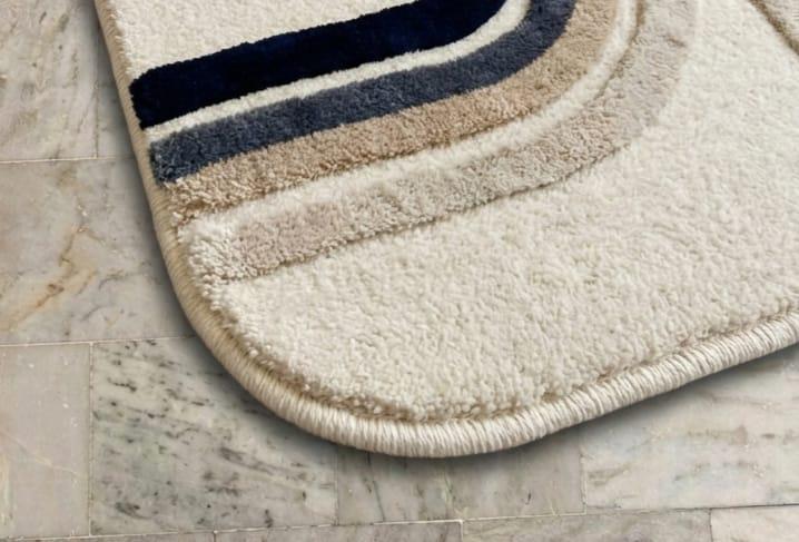 Detailed-hand-carved-high-low-pile-texture-geometric-archway-navy-blue-wool-rug  Close-up of a plush bath mat corner on a light marble tile floor, featuring concentric curved stripes in cream, beige, taupe, and dark navy.