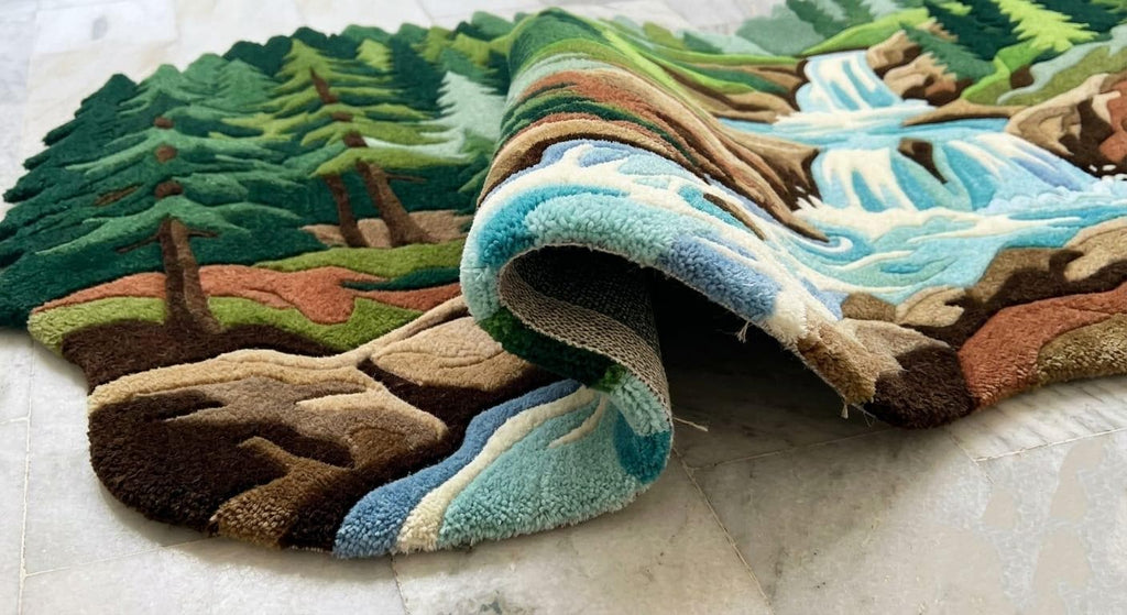  Shubham-Art-Rugs-hand-tufted-new-zealand-wool-forest-waterfall-nature-landscape-area-rug-rectangular A close-up of a textured, raised-loop area rug with a scenic landscape design featuring green pine trees, brown hills, and a blue and white river or waterfall, shown partially rolled over on a light-colored floor.