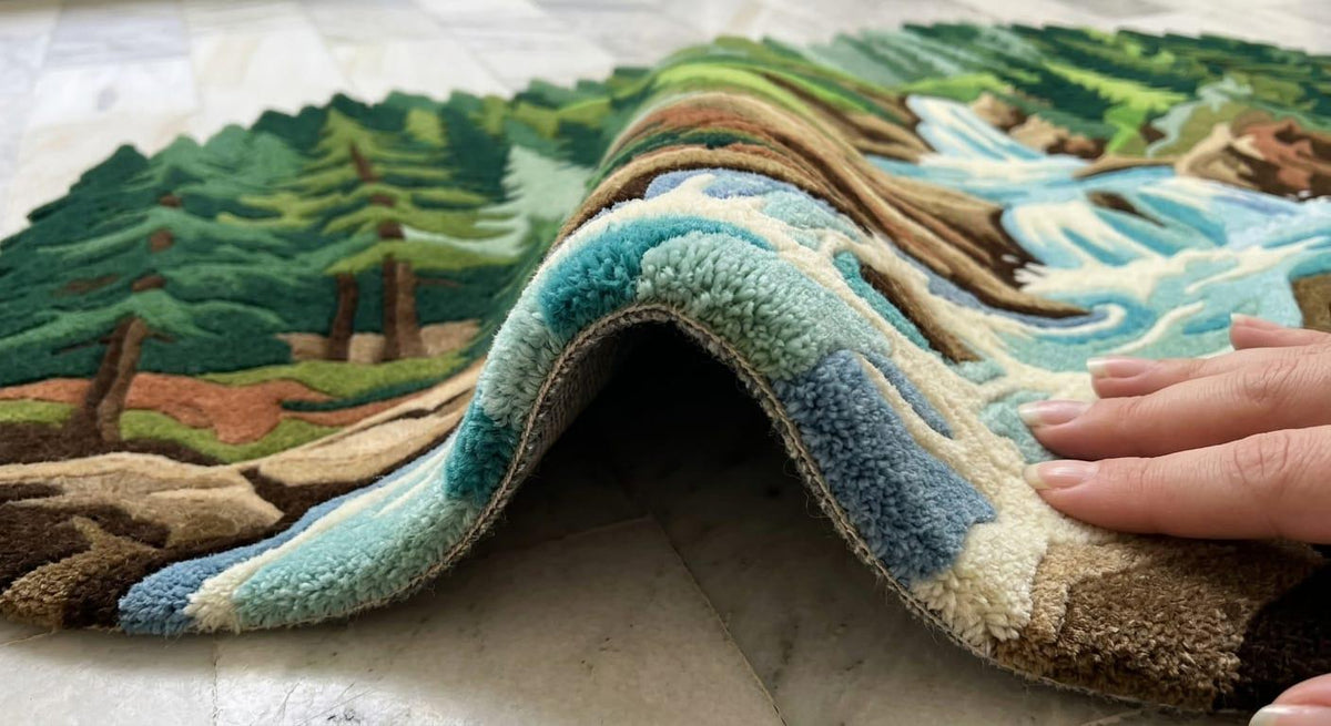  Modern-rustic-cabin-living-room-interior-decorated-with-large-rectangular-green-forest-waterfall-wool-carpet A close-up of a hand lifting a plush, multi-colored rug to reveal its layered landscape design, featuring green pine trees, brown hills, and blue waterways.