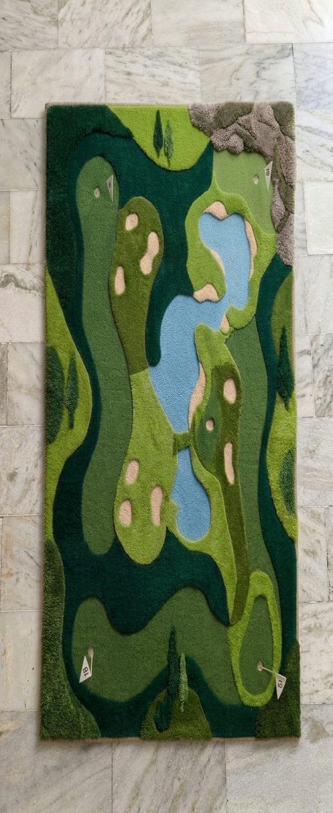 Shubham-Art-Rugs-hand-tufted-new-zealand-wool-golf-course-rug-novelty-sports-area-decorModern-game-room-office-interior-styling-featuring-luxury-shaped-golf-course-wool-carpet A rectangular wall-mounted rug featuring an abstract golf course landscape design in shades of green, dark green, light blue, and beige, displayed on a light gray marble tile wall.