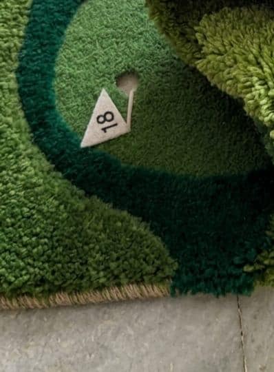 Detailed-3d-topography-hand-carved-high-low-pile-texture-sports-green-golf-rugModern-game-room-office-interior-styling-featuring-luxury-shaped-golf-course-wool-carpet Close-up of a green shag rug with a golf hole design, featuring a small white triangular tag marked '18' placed on the turf, with a gray floor visible at the bottom.