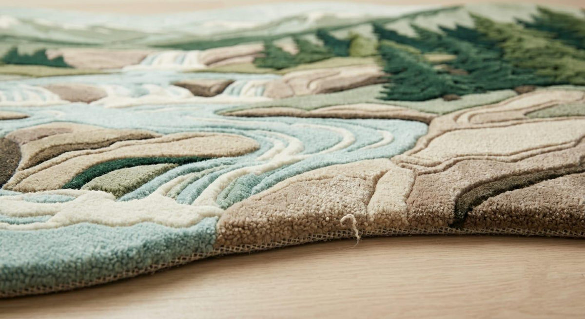 Detailed-hand-carved-high-low-pile-texture-new-zealand-wool-green-forest-beige-mountain-rug  A close-up of a textured area rug with a 3D mountain and river landscape design, featuring shades of beige, green, and blue, laid out on a light wood floor.