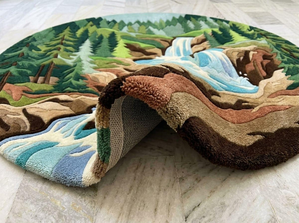  Circular-woodland-scenic-area-rug-3d-high-low-sculpted-pile-texture-forest-green-cascading-blue-waterfall A round, high-pile textured rug featuring a detailed landscape design of a waterfall flowing through a pine forest, shown partially folded over on a light-colored marble floor.