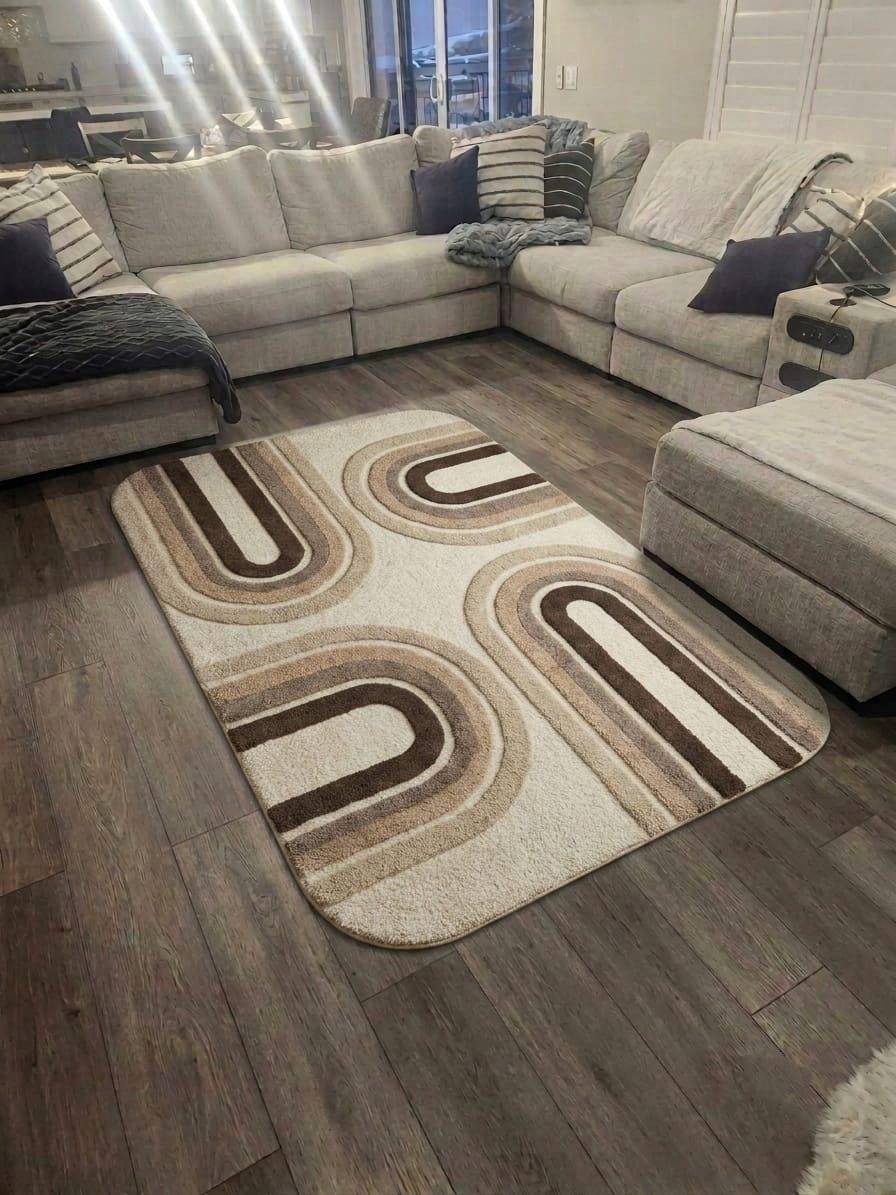 A rectangular area rug with a beige, brown, and cream geometric arch pattern, shown on a dark wood floor in a living room setting with a light gray sectional sofa.Shubham-Art-Rugs-hand-tufted-wool-arch-rug-mid-century-modern-neutral-high-low-pile-carpet