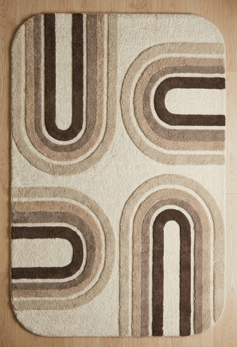 Shubham-Art-Rugs-hand-tufted-wool-arch-rug-mid-century-modern-neutral-high-low-pile-carpet A rectangular bath mat with rounded corners featuring a retro 1970s-style geometric pattern of concentric arches in beige, brown, and cream, laid flat on a light wood floor.