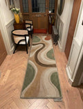 Shubham-Art-Rugs-hand-tufted-wool-area-rug-abstract-wavy-sage-green-terracotta-beige-contemporary-home-carpet A long rectangular area rug with an abstract wavy pattern in beige, brown, green, and cream, shown on a light wood floor in a hallway with wooden doors and chairs.
