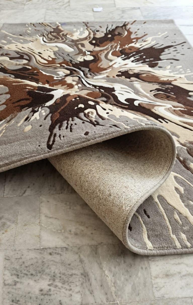 Shubham-Art-Rugs-hand-tufted-wool-rug-abstract-splash-design-contemporary-living-room-carpet  An abstract-patterned area rug with a rolled corner, displayed on a gray marble tile floor. The rug features a marbled design in shades of brown, beige, and cream, with a solid beige backing visible on the underside.