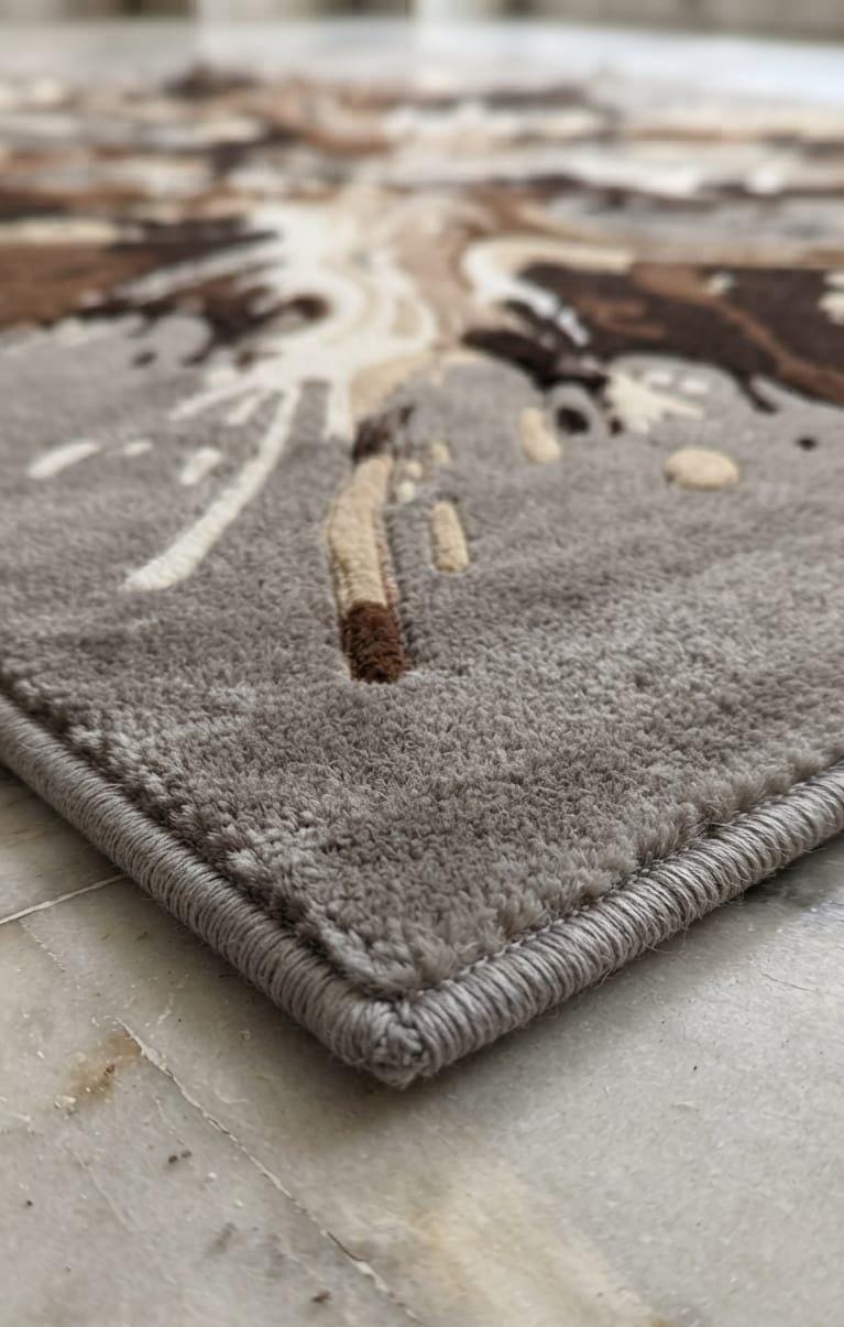 Detailed-hand-carved-3d-embossing-high-low-pile-texture-neutral-grey-mocha-brown-wool-rug  Close-up of a textured area rug corner showing a looped pile and braided edge, with an abstract pattern in taupe, cream, and brown on a light stone floor.