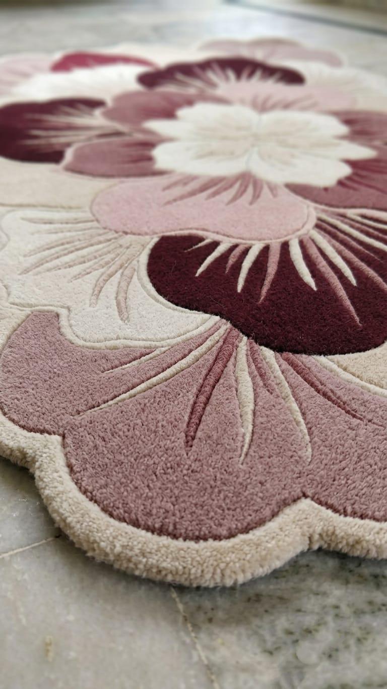 Detailed close up profile showing multi level sculpted pile texture on a handmade pink floral botanical wool rug
 A close-up of a floral-shaped rug with a raised, tufted pattern in dusty pink, cream, and deep burgundy, shown on a light stone floor.