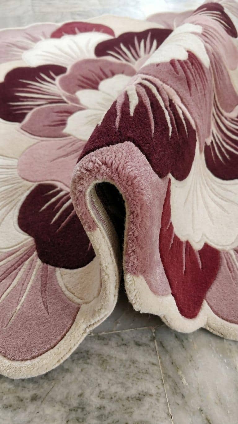 Detailed close up profile showing multi level sculpted pile texture on a handmade pink floral botanical wool rug
 A close-up of a plush area rug with a large-scale floral pattern in dusty pink, burgundy, and cream, draped over a gray marble floor.