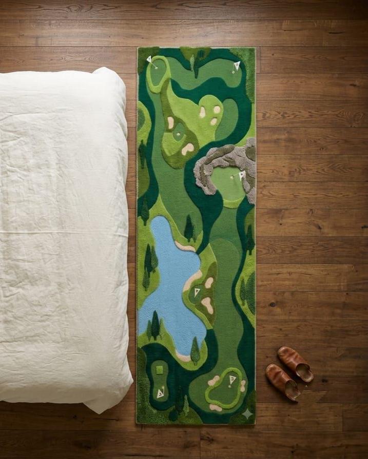 Shubham Art Rugs hand tufted New Zealand wool golf course runner rug featuring a 3D green fairway design layout A long rectangular area rug with a golf course landscape design in shades of green, blue, and beige, styled on a dark wood floor next to a white pillow and a pair of brown shoes.