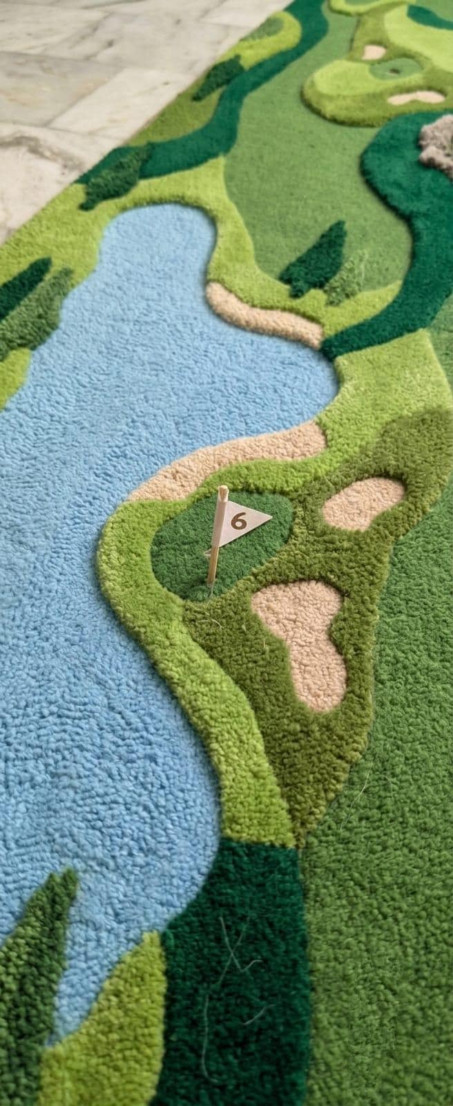Detailed close up profile of texturized multi level pile depths showing three dimensional fairway details A close-up of a textured, high-pile area rug shaped like a golf course, featuring green fairways and greens, light blue water features, and beige sand bunkers. A small white flag with the number 6 is placed on one of the greens.