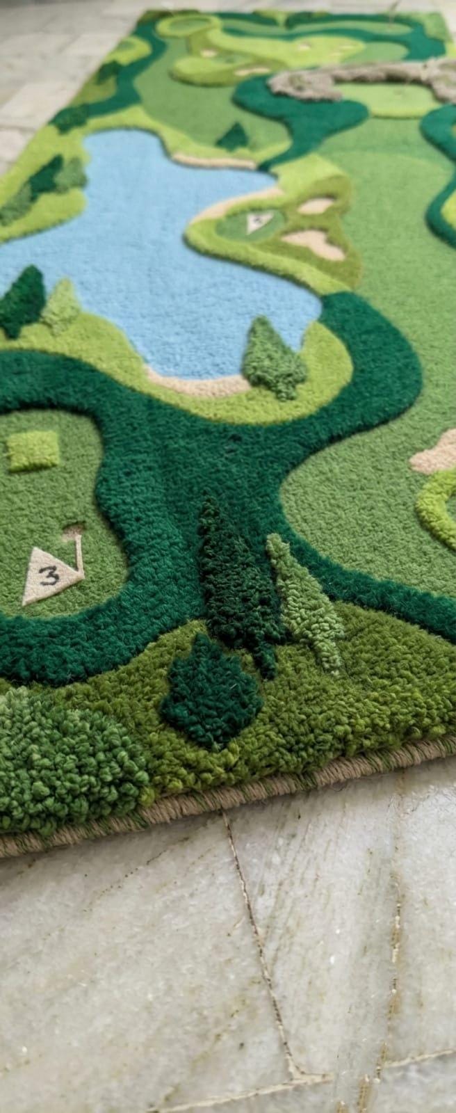 Detailed close up profile of texturized multi level pile depths showing three dimensional fairway details A close-up of a textured, golf-course-inspired area rug featuring green fairways, trees, and a light blue water feature, laid out on a light stone floor.