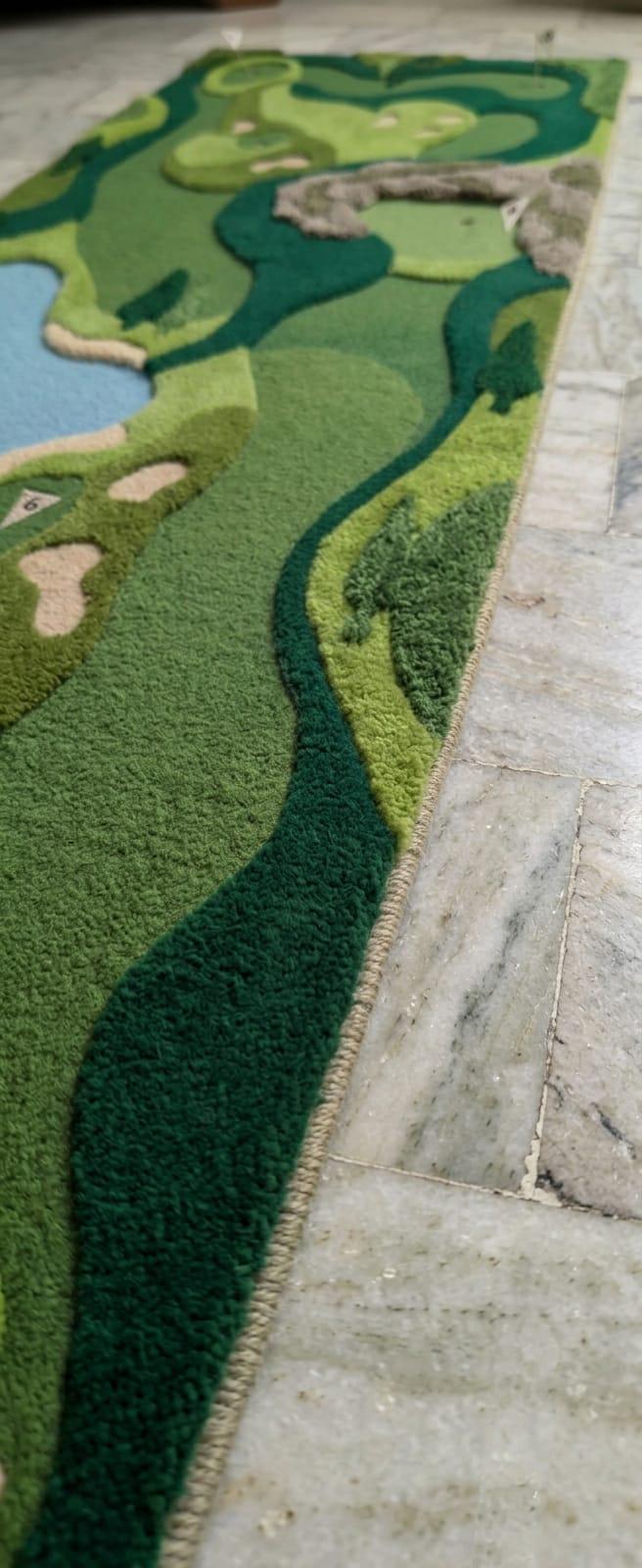 Luxury golf inspired floor runner rug styled in a modern residential hallway A long, low-pile area rug with a golf course landscape design in shades of green, dark green, beige, and blue, shown on a stone tile floor.