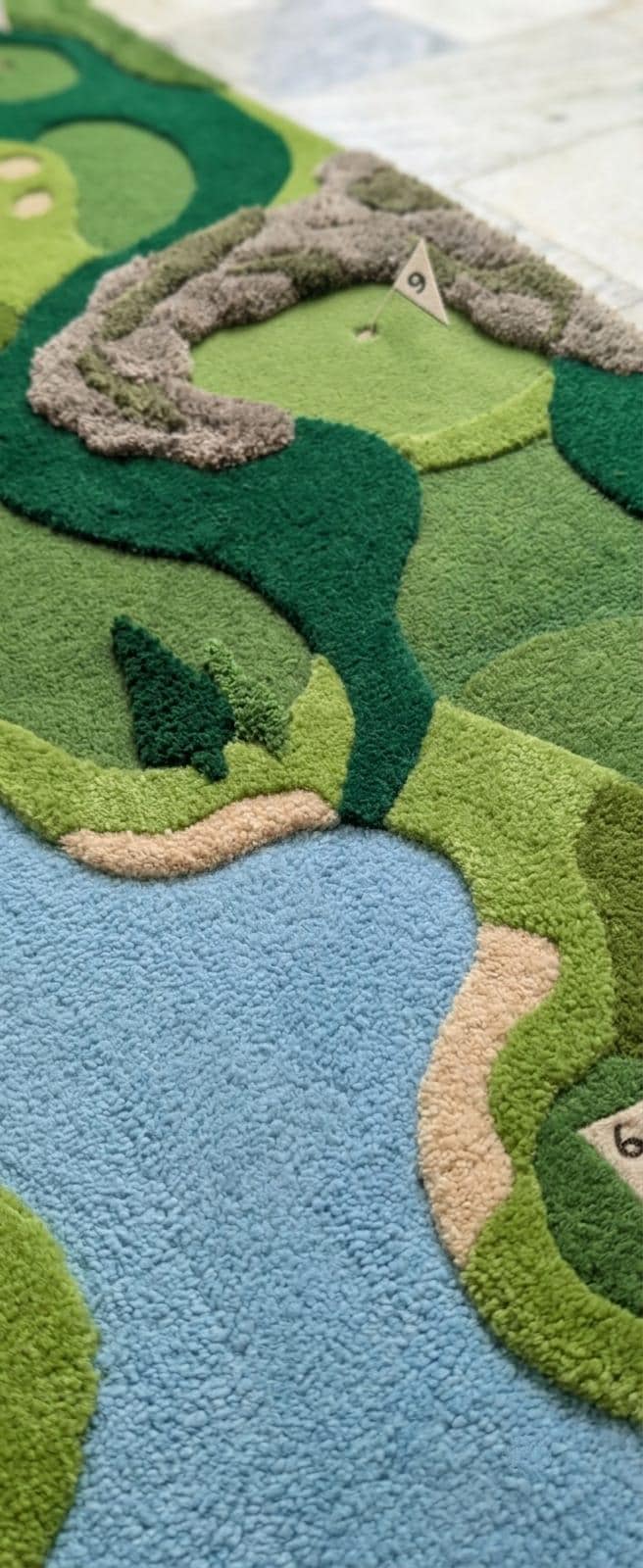 Detailed close up profile of texturized multi level pile depths showing three dimensional fairway details A close-up of a textured, raised rug shaped like a golf course landscape, featuring green and light green fairways, a blue water feature, and tan sand bunkers.