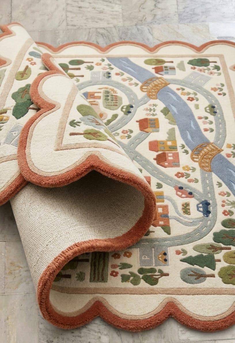 Detailed close up view of the dense plush wool pile detailing colorful little houses roads and trees on a premium handmade nursery carpet
 A close-up of a decorative area rug with a cream base and a whimsical country road scene. The rug features a scalloped terracotta border, a winding blue river, small houses, trees, and cars in muted green, orange, and brown tones.