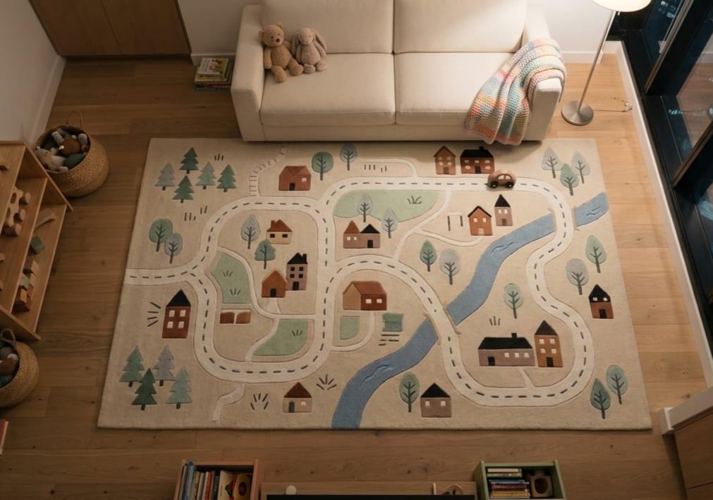Shubham Art Rugs hand tufted wool city map play rug featuring a boho neutral nursery road layout A large rectangular children's play rug featuring a whimsical village road design with houses, trees, and a winding blue river, shown on a wooden floor in a styled living room setting.